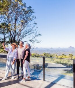 Three women are taking a picture on a balcony overlooking the mountains during a Sunshine Coast tour.