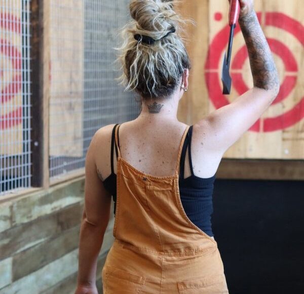 A woman in overalls holding a bow and arrow in front of a target during a sunshine coast day trip.