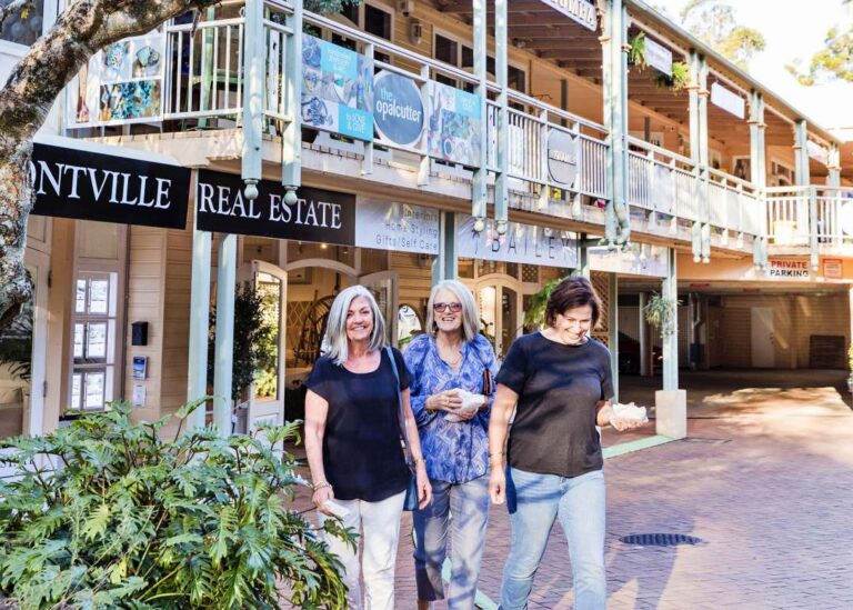 Three individuals strolling and smiling in front of a two-story building with signage for Montville Real Estate during their Montville Day Tour. Sunshine Coast Hinterland Highlights Tour: Discover Maleny and Montville