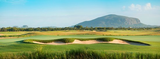 A panoramic view of a golf course with manicured greens and sand bunkers in the foreground, with a large, rounded mountain rising in the background under a clear sky on the Sunshine Coast.